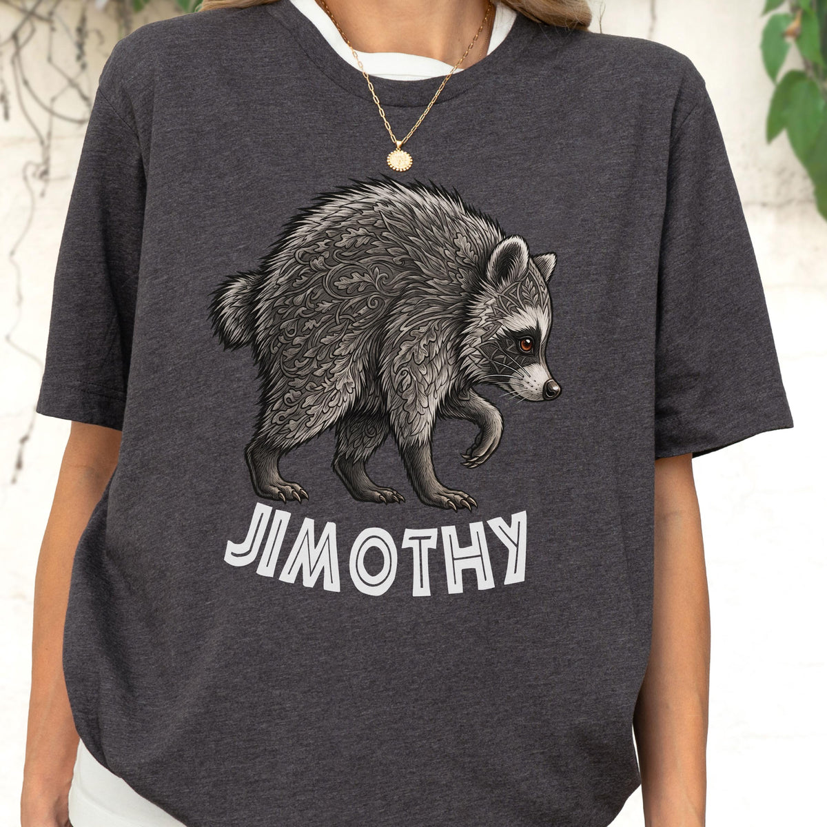 May include: A dark charcoal gray t-shirt featuring a detailed illustration of a raccoon with intricate leaf-like patterns on its fur. Below the raccoon, the text "JIMOTHY" is printed in a bold, white, sans-serif font. The shirt has short sleeves.
