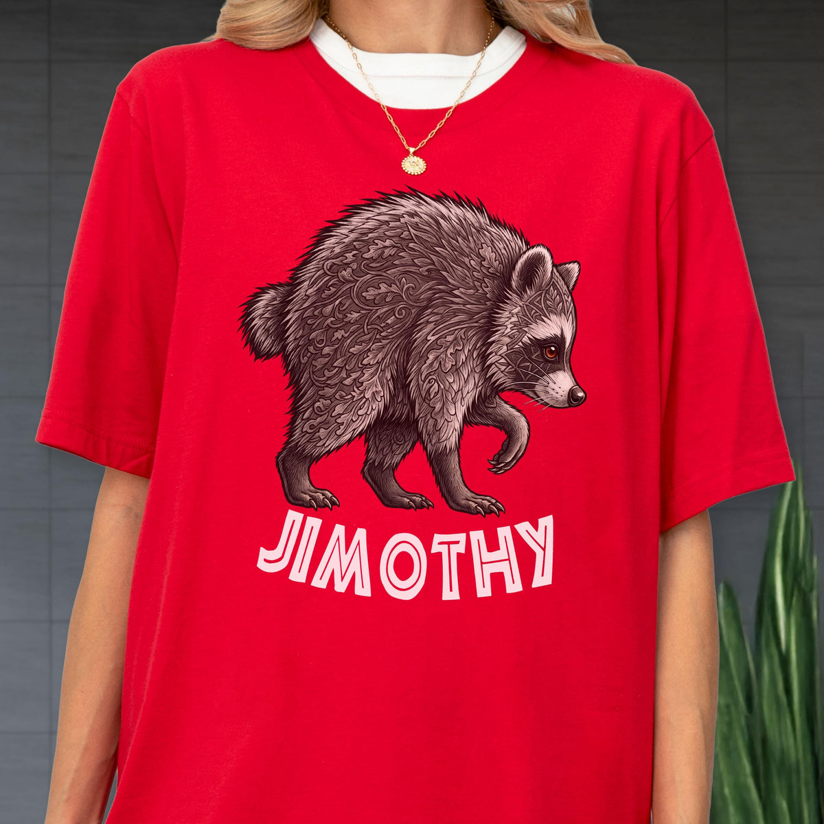 May include: A bright red t-shirt featuring a graphic illustration of a raccoon with intricate leaf-like patterns in its fur. Below the raccoon, the name JIMOTHY is printed in bold, white, sans-serif capital letters. The shirt has a crew neckline.