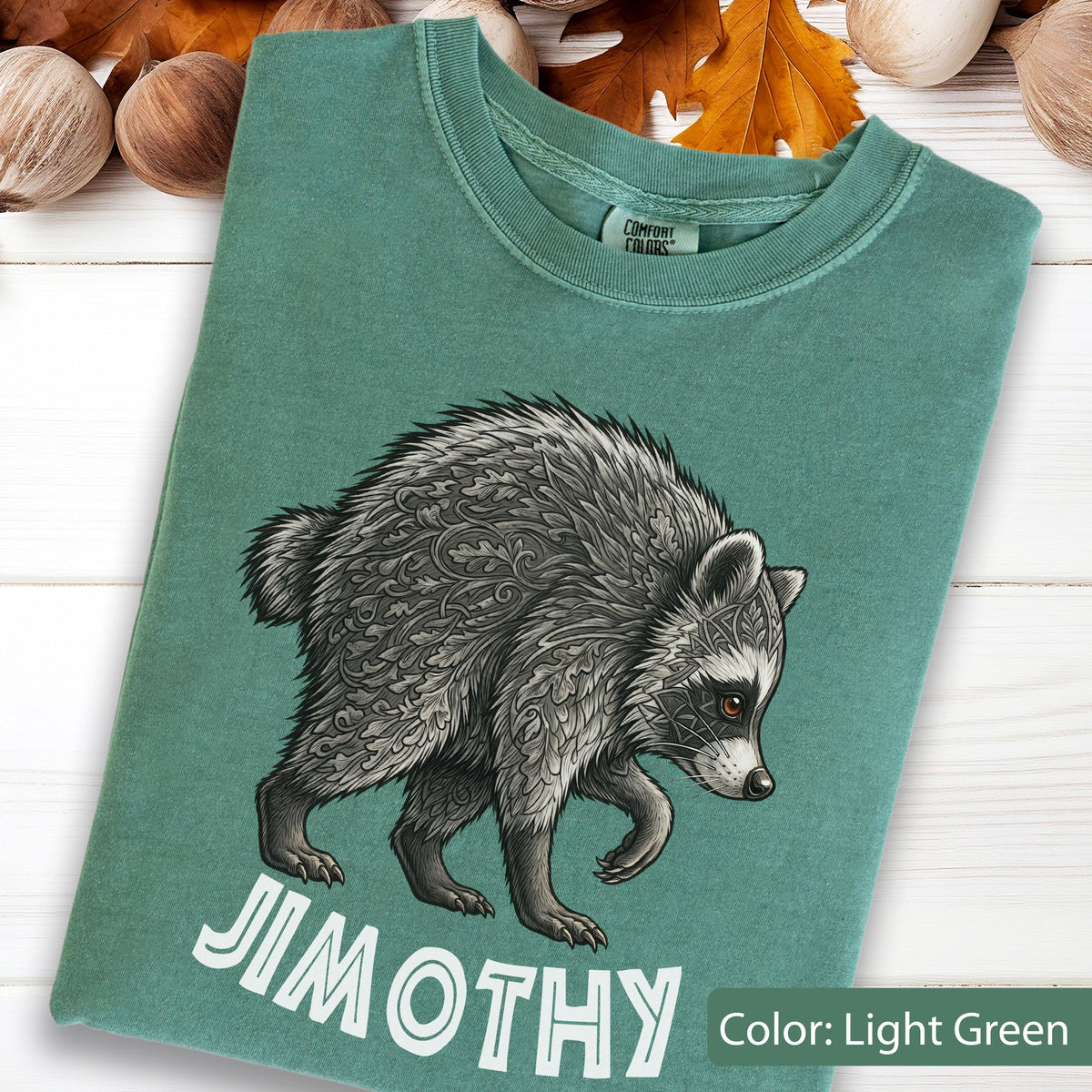 May include: A light green crew neck t-shirt featuring a graphic of a raccoon with intricate leaf-patterned fur. The raccoon is depicted in shades of gray and black. Below the animal, the text JIMOTHY is printed in large, bold, white capital letters.