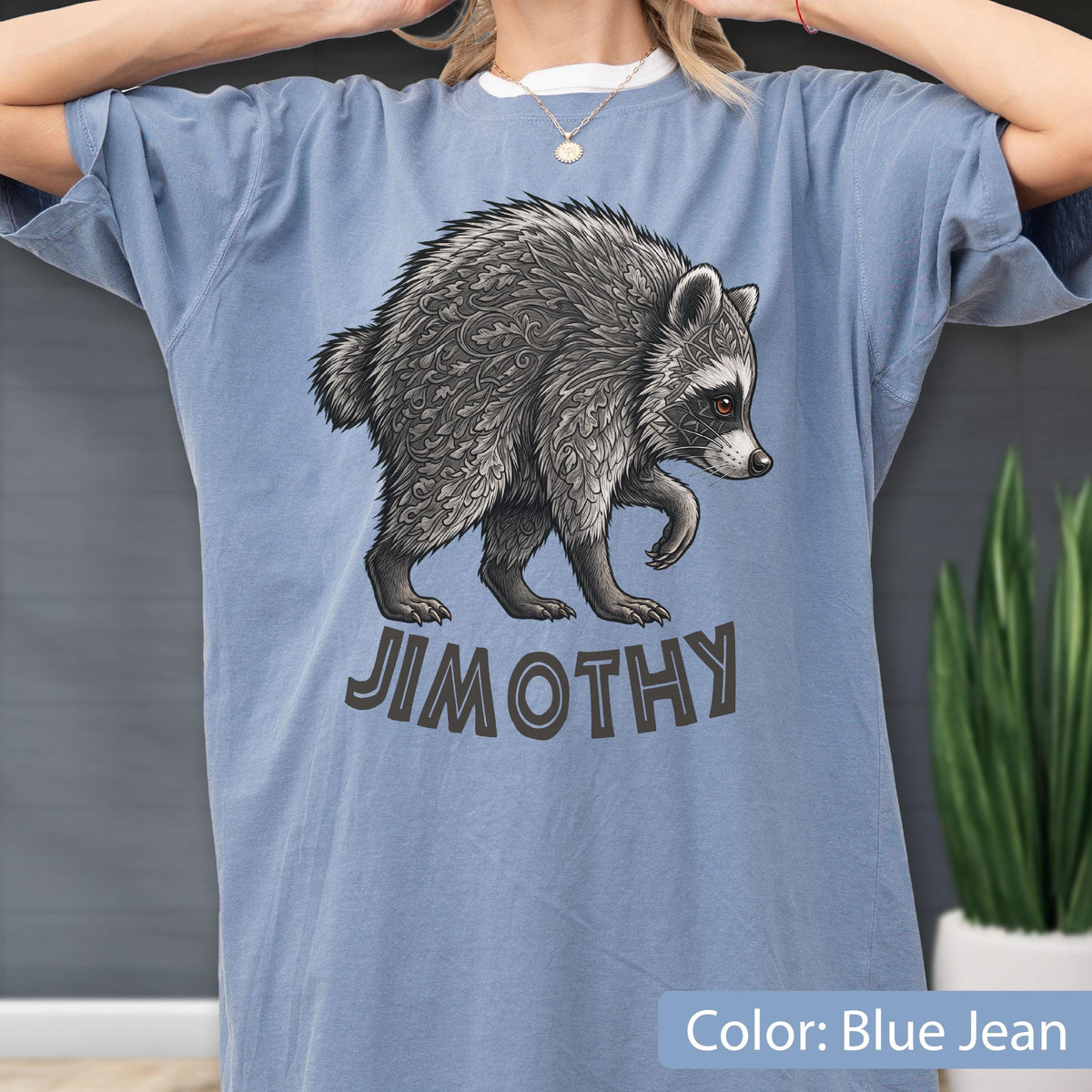 May include: A blue jean colored t-shirt featuring a graphic illustration of a raccoon with intricate leaf-like patterns in its fur. Below the raccoon, the text JIMOTHY is printed in a bold, dark font. The shirt has short sleeves and a crew neckline.