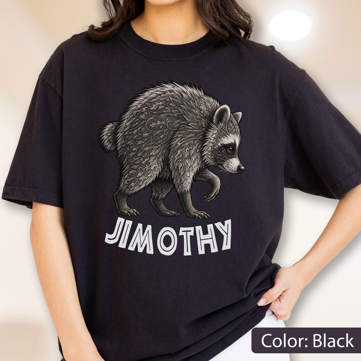 May include: A black short-sleeved t-shirt featuring a graphic of a raccoon with intricate grey leaf-like patterns on its fur. Below the raccoon, the text JIMOTHY is printed in bold, white, capital letters with a distinct outlined font style.