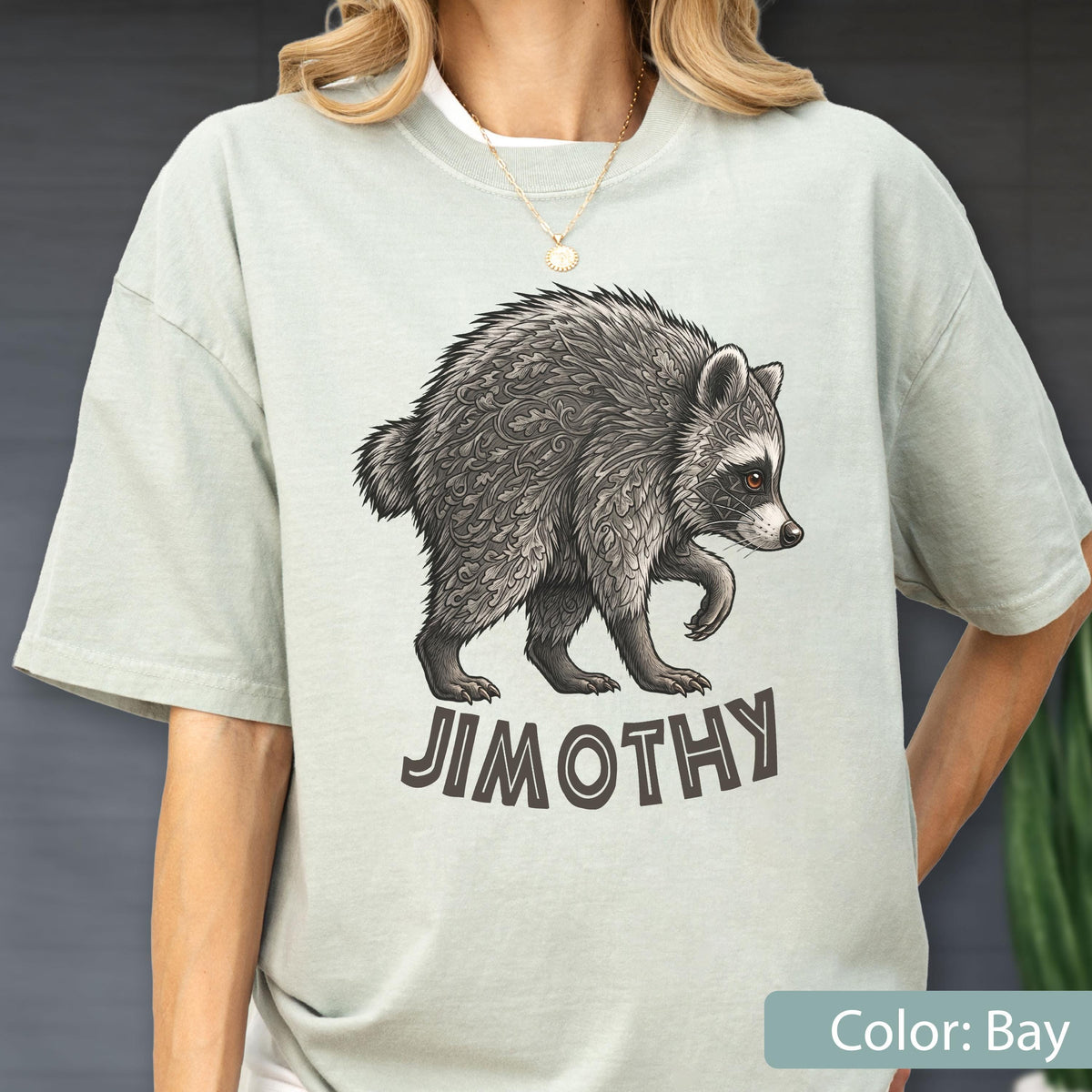 May include: A light sage green short-sleeve t-shirt featuring a detailed illustration of a raccoon with intricate fur patterns. Below the raccoon, the name JIMOTHY is printed in bold, dark brown capital letters. The shirt has a crew neckline.