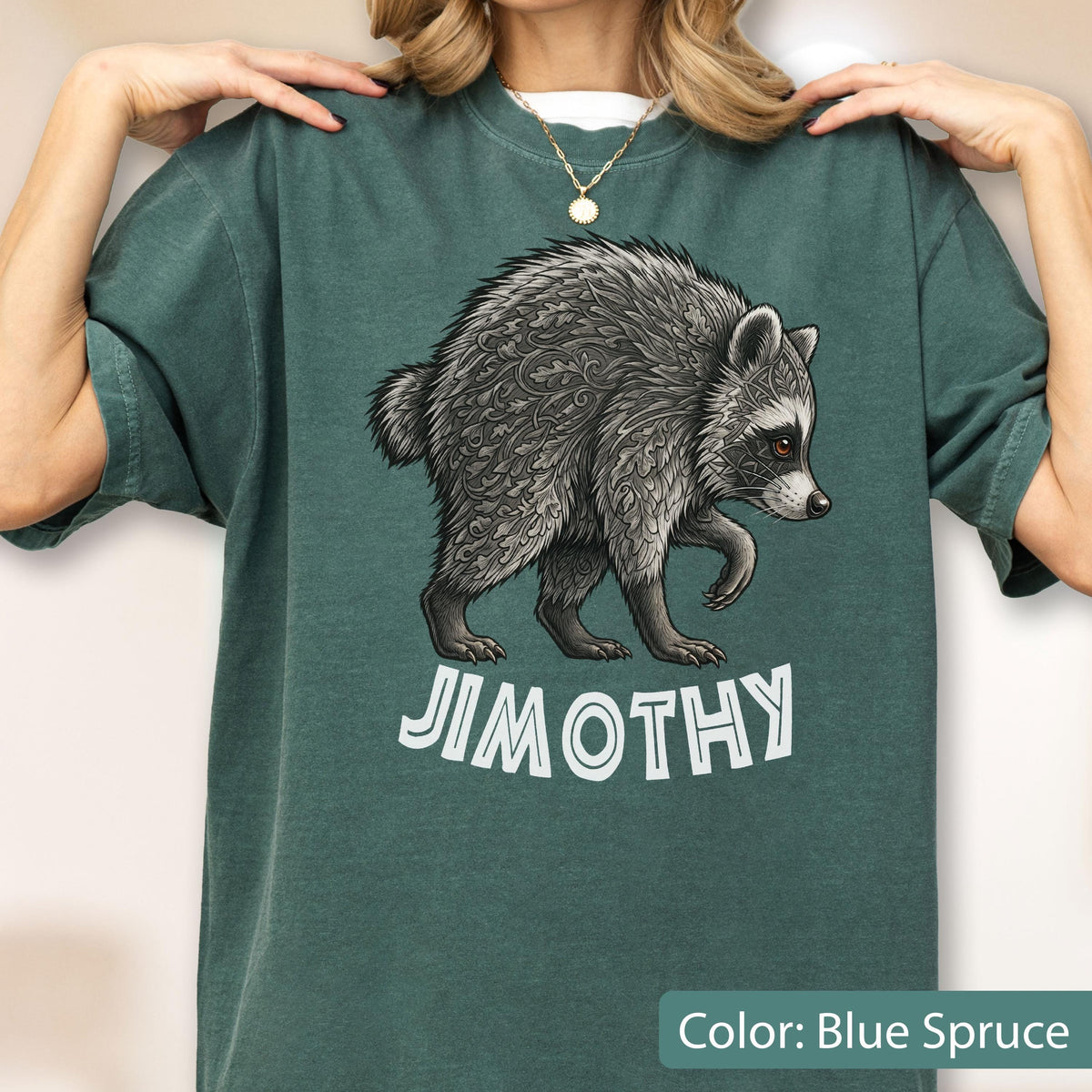 May include: A blue spruce short-sleeve t-shirt featuring a graphic of a raccoon with intricate leaf-like patterns in its fur. Below the raccoon, the text JIMOTHY is printed in a bold, white, sans-serif font. The shirt has a crew neckline.