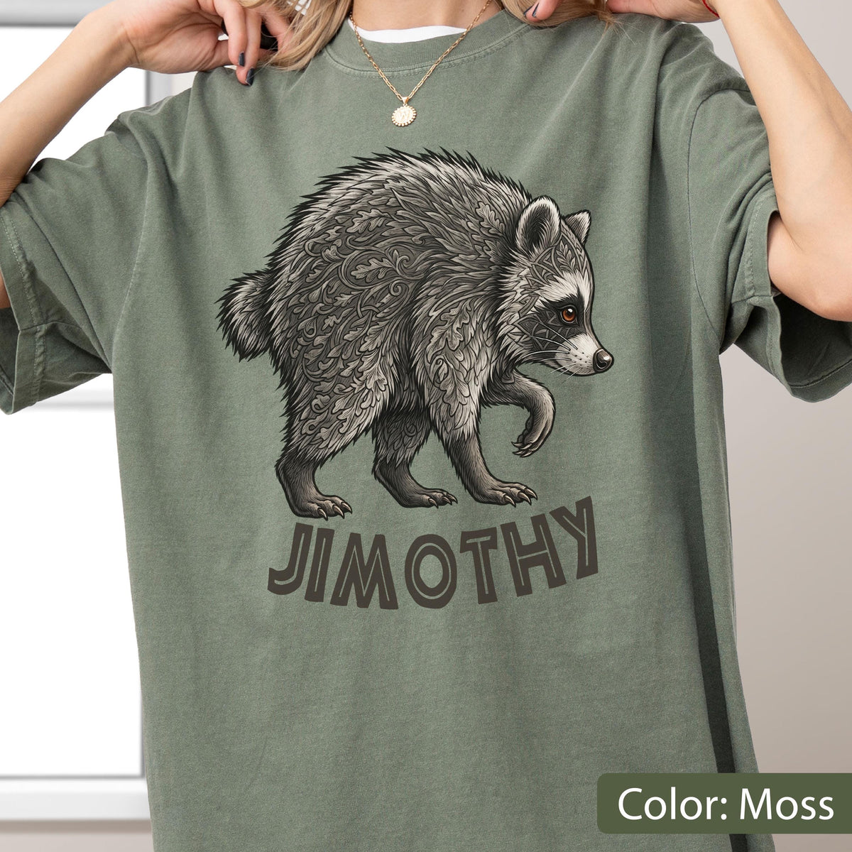 May include: A moss green t-shirt featuring a graphic illustration of a raccoon with intricate leaf-like patterns in its fur. Below the raccoon, the name JIMOTHY is printed in bold, dark brown capital letters. The shirt has a crew neckline.