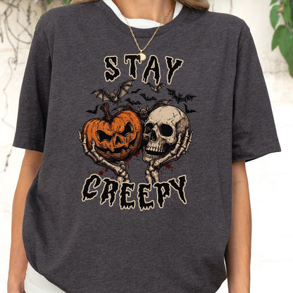 May include: A dark heather gray t-shirt featuring a graphic print of two skeletal hands holding an orange jack-o'-lantern and a human skull. Above and below the illustration, the text reads STAY CREEPY in a jagged, black, horror-style font.