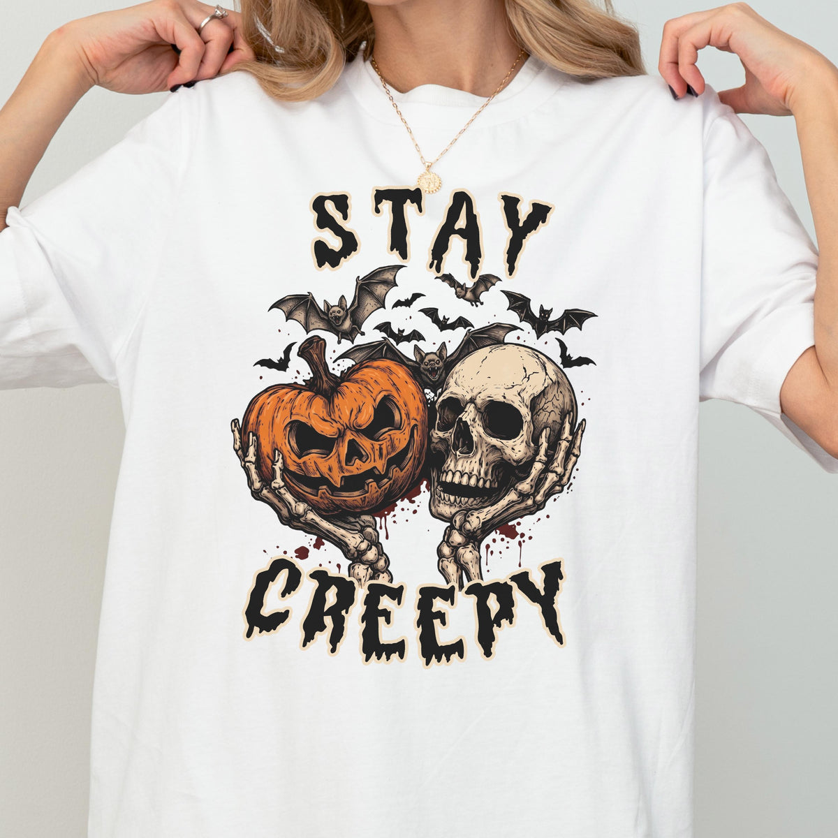 May include: A white t-shirt featuring a graphic print of two skeletal hands holding an orange jack-o'-lantern and a human skull. Above and below the illustration, the text reads STAY CREEPY in a black, jagged font, surrounded by small black bats.