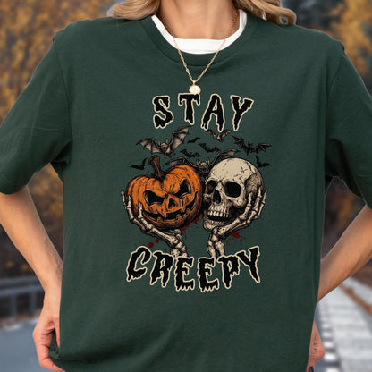 May include: A dark green t-shirt featuring a graphic print of two skeletal hands holding an orange jack-o'-lantern and a human skull. Above and below the central illustration, the text reads STAY CREEPY in a stylized, dripping black font.