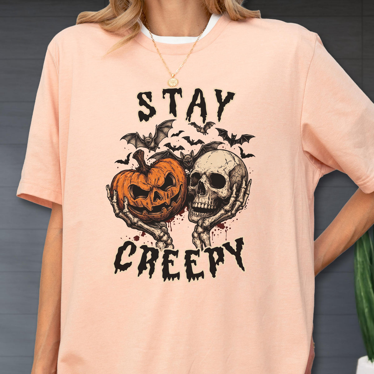 May include: A peach-colored short-sleeve t-shirt featuring a graphic print of a jack-o'-lantern and a human skull held by skeletal hands. Above and below the illustration, the text reads STAY CREEPY in a black, jagged, distressed font.