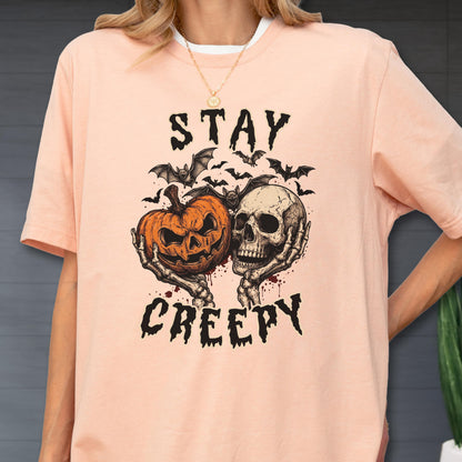 May include: A peach-colored short-sleeve t-shirt featuring a graphic print of a jack-o'-lantern and a human skull held by skeletal hands. Above and below the illustration, the text reads STAY CREEPY in a black, jagged, distressed font.