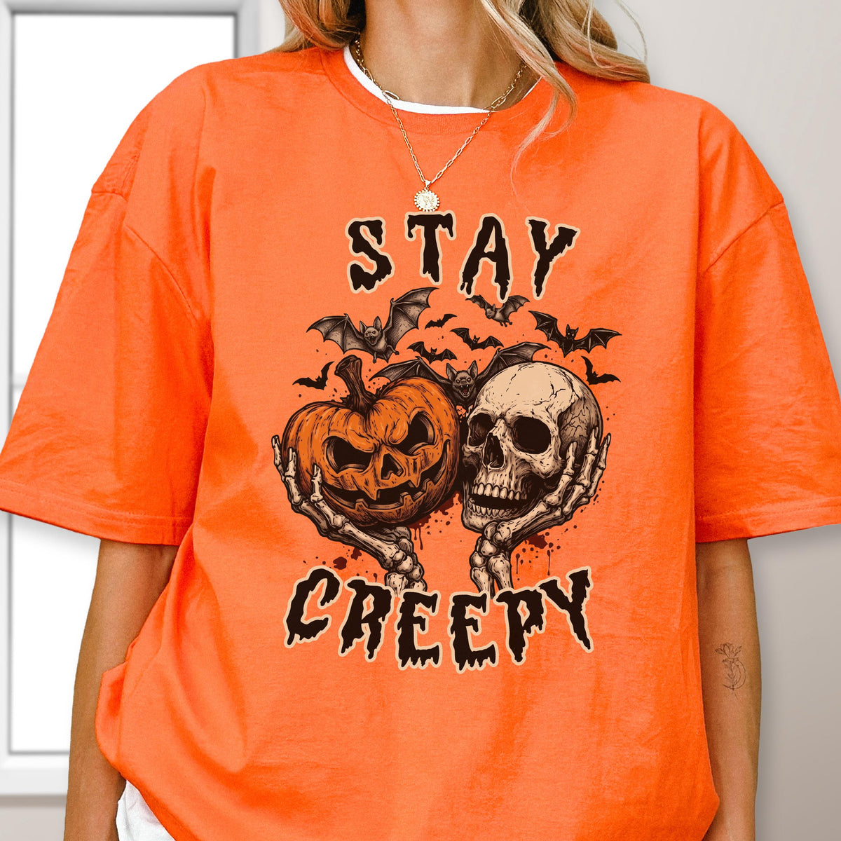 May include: An orange short-sleeve t-shirt featuring a graphic print of a jack-o'-lantern and a human skull held by skeletal hands, surrounded by bats. The text "STAY CREEPY" is printed in a dark, jagged font above and below the illustration.