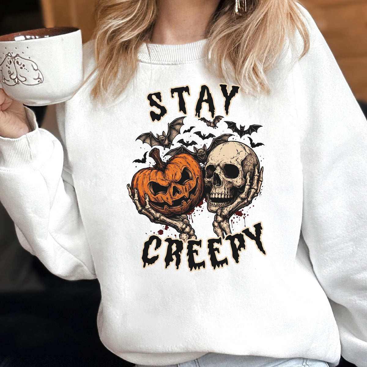 May include: A white crewneck sweatshirt featuring a graphic print of a jack-o'-lantern and a human skull held by skeletal hands, surrounded by bats. The text "STAY CREEPY" is printed in a black, jagged font above and below the central illustration.