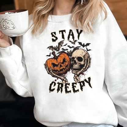 May include: A white crewneck sweatshirt featuring a graphic print of a jack-o'-lantern and a human skull held by skeletal hands, surrounded by bats. The text "STAY CREEPY" is printed in a black, jagged font above and below the central illustration.