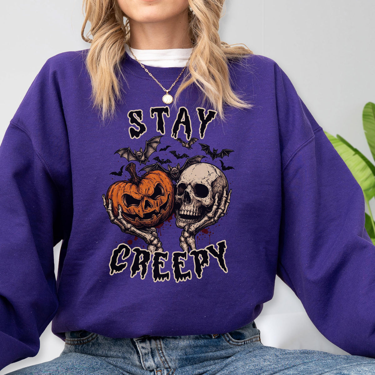 May include: A purple long-sleeve sweatshirt featuring a graphic print of a jack-o'-lantern and a human skull held by skeletal hands. Above and below the central image, the text reads STAY and CREEPY in a jagged, dark font, surrounded by bats.