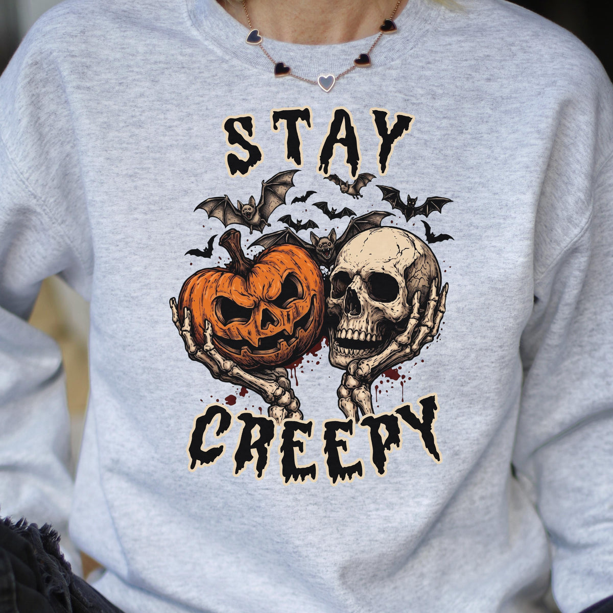 May include: A light gray crewneck sweatshirt featuring a graphic print of a jack-o'-lantern and a human skull held by skeletal hands. Above and below the central illustration, the text reads STAY and CREEPY in a jagged, black font.