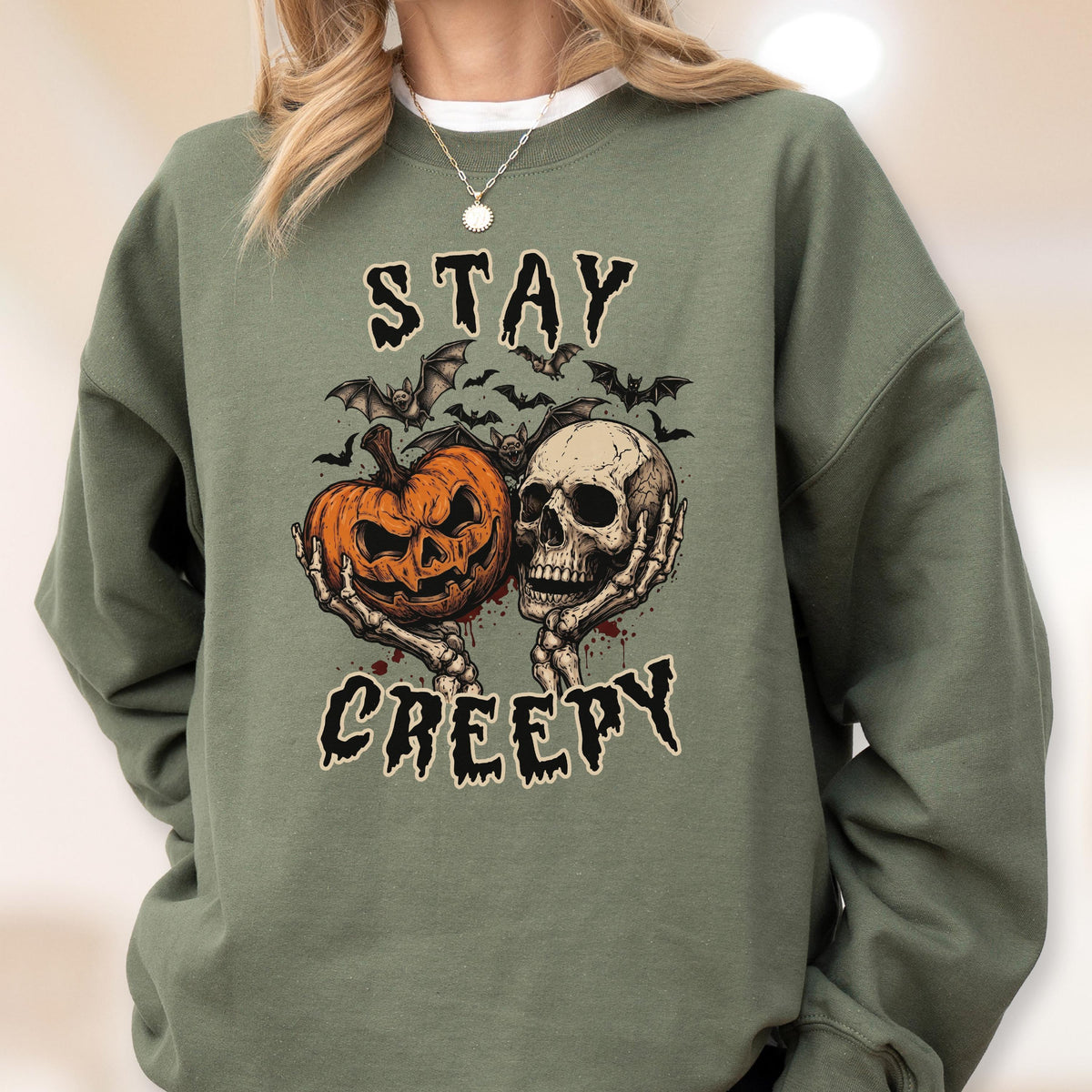 May include: An olive green crewneck sweatshirt featuring a graphic print of a jack-o'-lantern and a human skull held by skeletal hands. Above and below the illustration, the text reads STAY CREEPY in a jagged, dark font, surrounded by small bats.