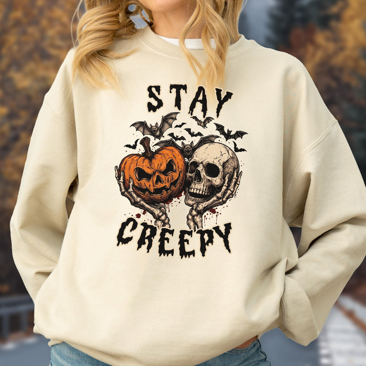 May include: A beige crewneck sweatshirt featuring a graphic print of a jack-o'-lantern and a human skull held by skeletal hands. Above and below the central illustration, the text reads STAY and CREEPY in a jagged, dark font with bats.