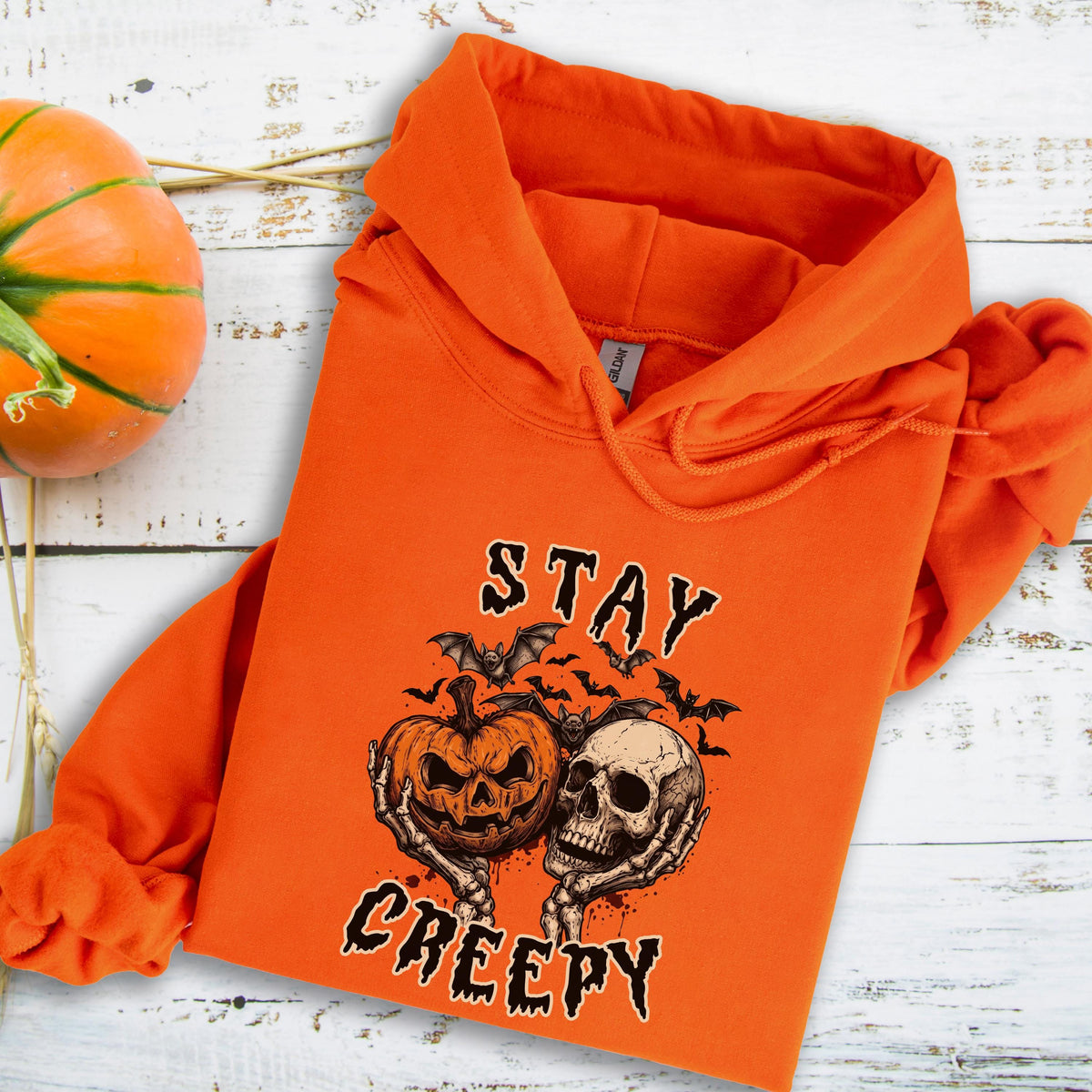 May include: An orange hooded sweatshirt featuring a graphic print of a jack-o'-lantern and a human skull held by skeletal hands, surrounded by bats. The text "STAY CREEPY" is printed in a stylized black font above and below the central illustration.
