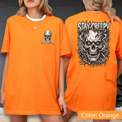 May include: An orange t-shirt featuring a small skull graphic and the text "STAY CREEPY" on the front left chest. The back of the shirt displays a large, detailed black and white illustration of a skull with intricate line work and the text "STAY CREEPY" above it.