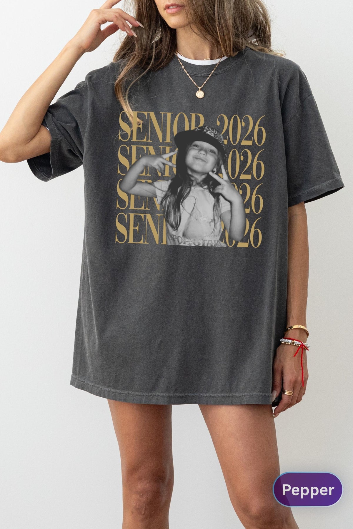 May include: A charcoal gray oversized t-shirt featuring a graphic print of a child wearing a baseball cap and making peace signs with both hands. The text SENIOR 2026 is repeated in gold lettering across the front, layered over the central image.