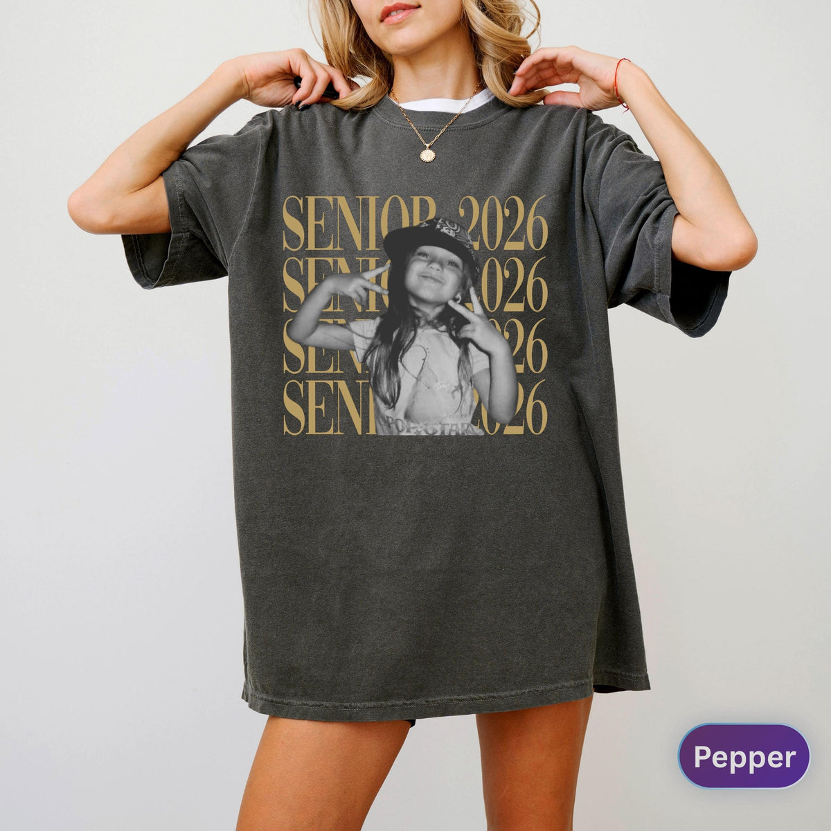 May include: A charcoal gray oversized t-shirt featuring a black and white photograph of a person wearing a cap and making a peace sign. The text SENIOR 2026 is printed in a large gold serif font repeatedly behind the central image.