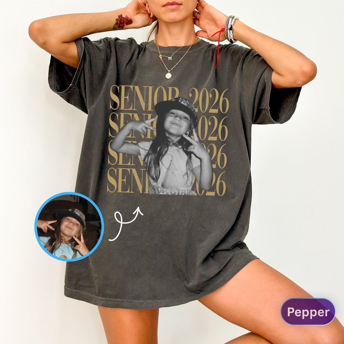 May include: A charcoal gray oversized t-shirt featuring a repeating gold text design that reads SENIOR 2026. The center graphic displays a black and white photograph of a person wearing a bucket hat and making a peace sign, with a circular inset.