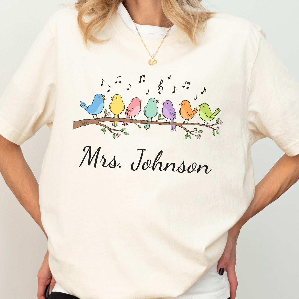 May include: A cream-colored short-sleeve t-shirt featuring a graphic of seven colorful birds perched on a brown branch with musical notes floating above them. Below the birds, the text Mrs. Johnson is printed in a black cursive script font.