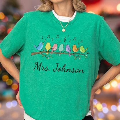 May include: A bright green short-sleeve t-shirt featuring a graphic of seven colorful birds perched on a brown tree branch with musical notes floating above them. The text "Mrs. Johnson" is printed in black cursive script below the branch.