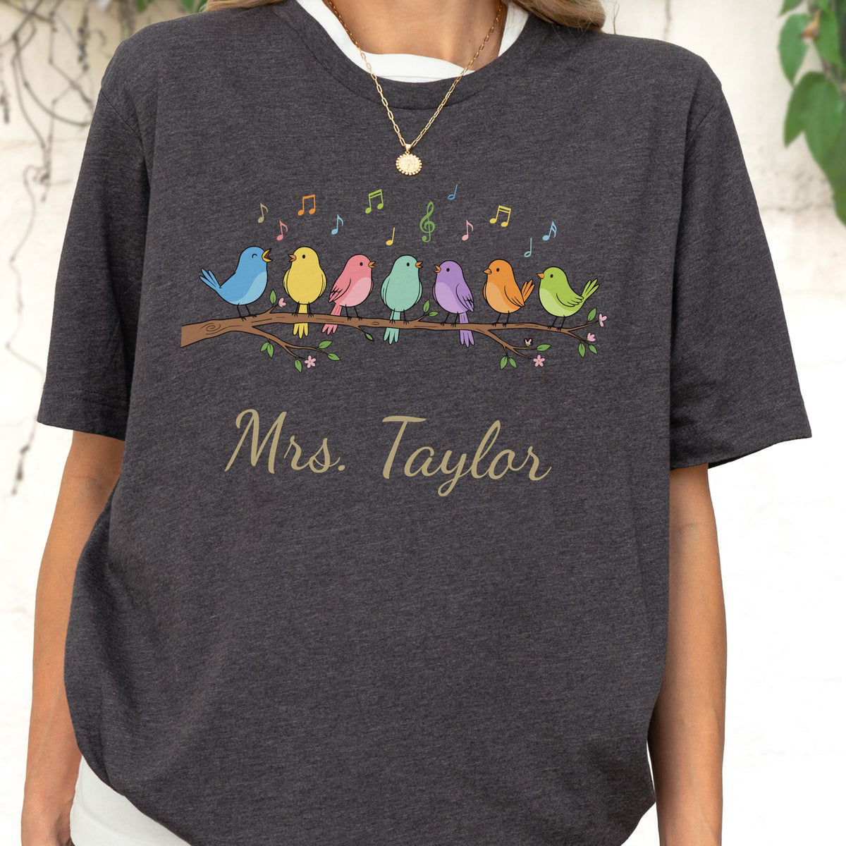 May include: A dark charcoal gray t-shirt featuring a graphic of seven colorful birds perched on a brown branch with musical notes floating above them. The text "Mrs. Taylor" is printed in a gold cursive font below the bird illustration.