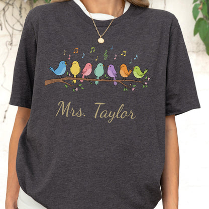 May include: A dark charcoal gray t-shirt featuring a graphic of seven colorful birds perched on a brown branch with musical notes floating above them. The text "Mrs. Taylor" is printed in a gold cursive font below the bird illustration.