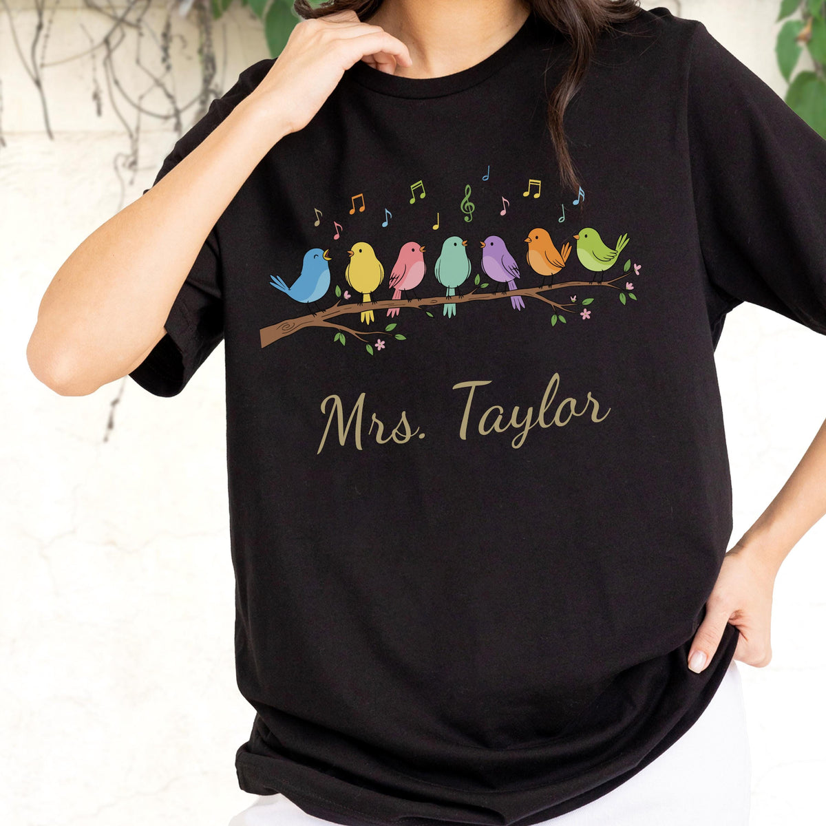 May include: A black short-sleeved t-shirt featuring a graphic of seven colorful birds perched on a brown tree branch with musical notes above them. Below the branch, the text "Mrs. Taylor" is printed in a gold cursive script font.