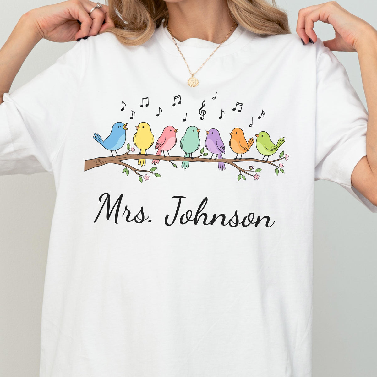 May include: A white t-shirt featuring a graphic of seven colorful birds perched on a brown branch with green leaves and small pink flowers. Above the birds are black musical notes. The text "Mrs. Johnson" is printed in black cursive script below.