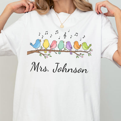May include: A white t-shirt featuring a graphic of seven colorful birds perched on a brown branch with green leaves and small pink flowers. Above the birds are black musical notes. The text "Mrs. Johnson" is printed in black cursive script below.