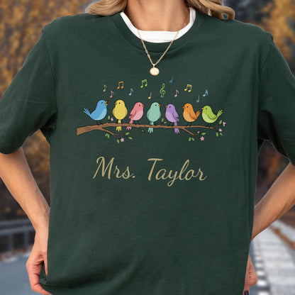 May include: A dark green t-shirt featuring a graphic of seven colorful birds perched on a brown branch with musical notes floating above them. The text "Mrs. Taylor" is printed in a gold script font below the branch illustration.
