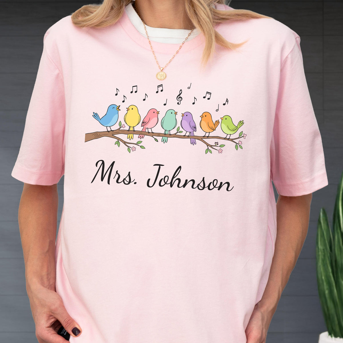 May include: A light pink t-shirt featuring a graphic of seven colorful birds perched on a brown branch with musical notes floating above them. Below the birds, the text Mrs. Johnson is printed in a black cursive script font on the front.