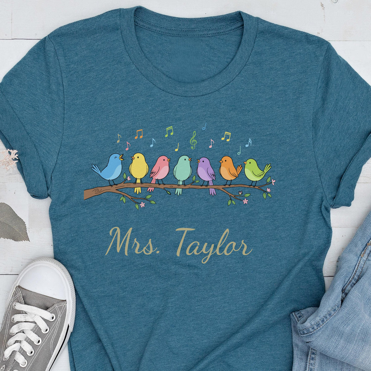 May include: A teal short-sleeved t-shirt featuring a graphic of seven colorful birds perched on a branch with musical notes above them. The text "Mrs. Taylor" is printed in a cursive font below the birds on the front of the garment.