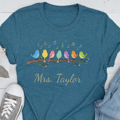 May include: A teal short-sleeved t-shirt featuring a graphic of seven colorful birds perched on a branch with musical notes above them. The text "Mrs. Taylor" is printed in a cursive font below the birds on the front of the garment.