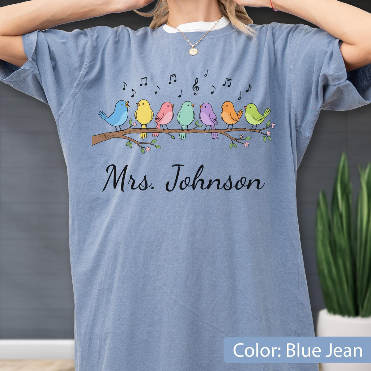 May include: A blue jean colored t-shirt featuring a graphic of seven colorful birds perched on a brown tree branch with musical notes above them. The text "Mrs. Johnson" is printed in black cursive script below the birds on the front of the shirt.