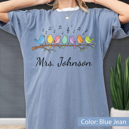 May include: A blue jean colored t-shirt featuring a graphic of seven colorful birds perched on a brown tree branch with musical notes above them. The text "Mrs. Johnson" is printed in black cursive script below the birds on the front of the shirt.
