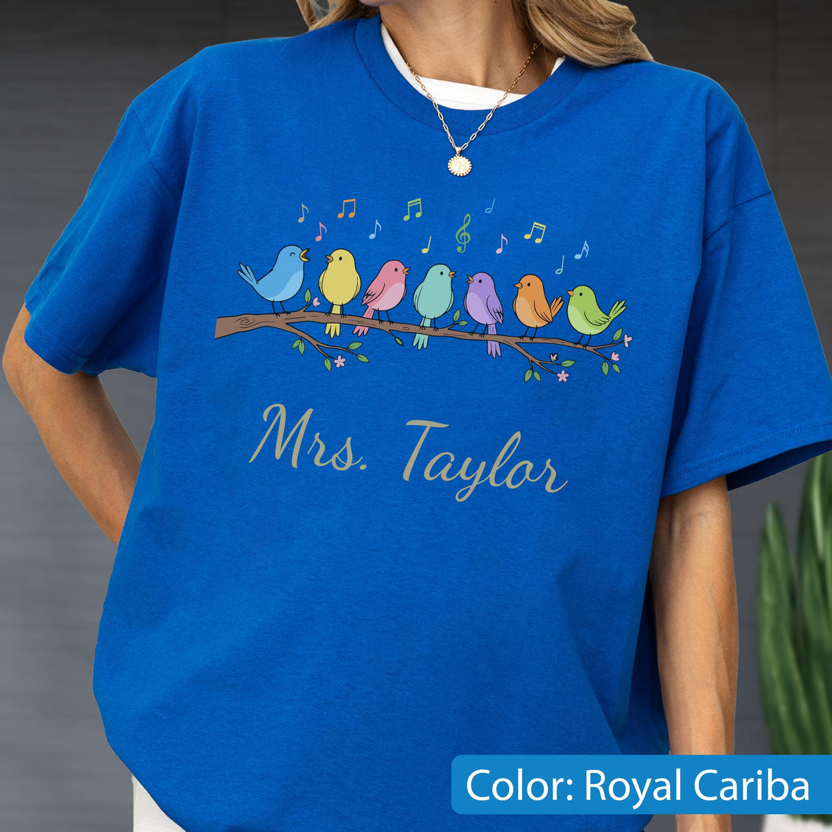 May include: A royal blue t-shirt featuring a graphic of seven colorful birds perched on a brown branch with musical notes floating above them. Below the birds, the text Mrs. Taylor is printed in a cursive script font in a light gray color.