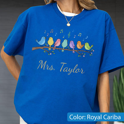 May include: A royal blue t-shirt featuring a graphic of seven colorful birds perched on a brown branch with musical notes floating above them. Below the birds, the text Mrs. Taylor is printed in a cursive script font in a light gray color.