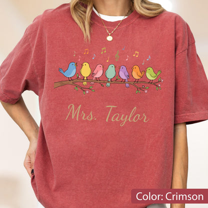 May include: A crimson short-sleeve t-shirt featuring a graphic of seven colorful birds perched on a brown branch with musical notes above them. The text "Mrs. Taylor" is printed in a cursive font below the birds. The shirt has a crew neckline.