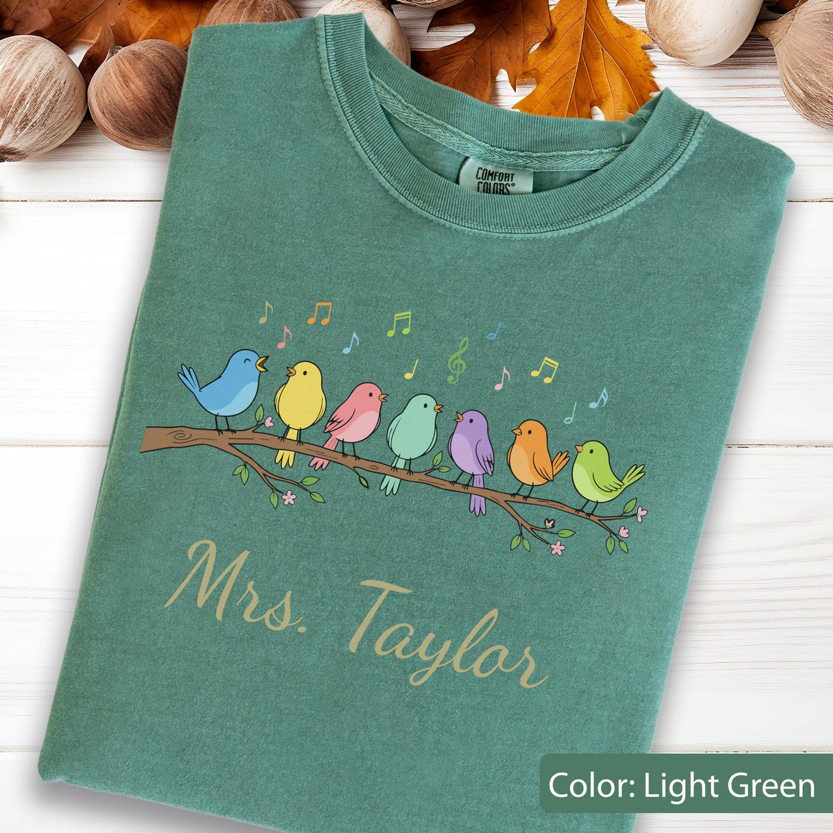 May include: A light green crewneck t-shirt featuring a graphic of seven colorful birds perched on a brown branch with musical notes above them. The text "Mrs. Taylor" is printed in a light tan script font below the bird illustration.