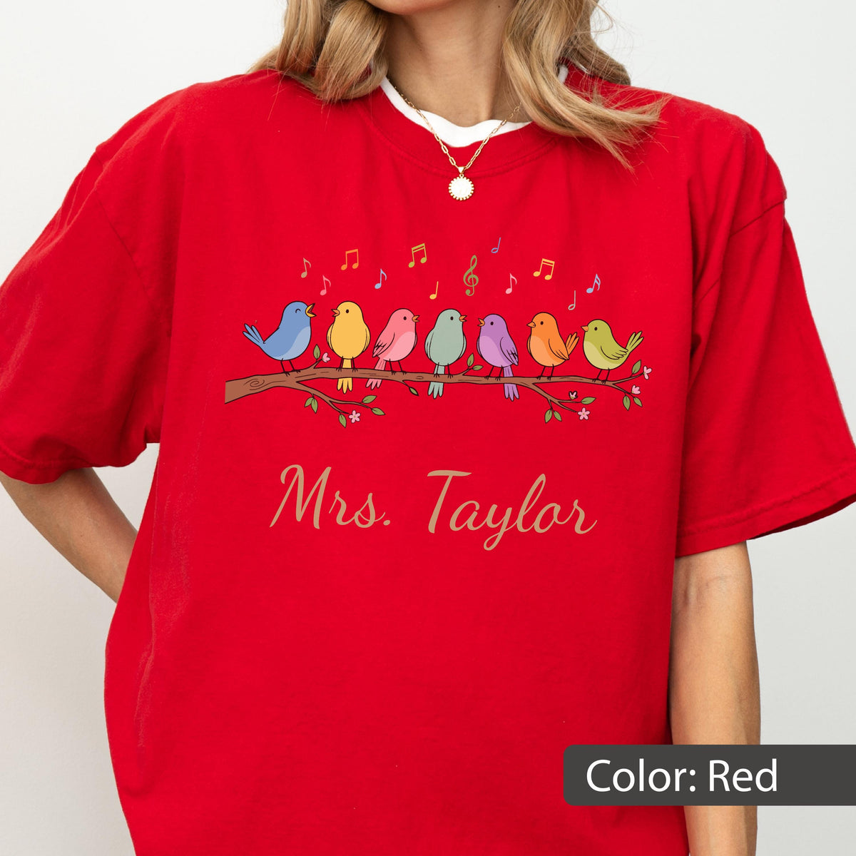 May include: A bright red short-sleeve t-shirt featuring a graphic of seven colorful birds perched on a brown branch. Above the birds are yellow and red musical notes. The text "Mrs. Taylor" is printed in a cursive gold font below the branch.