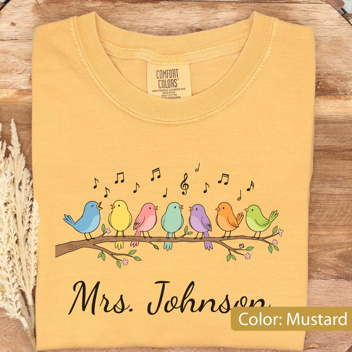 May include: A mustard yellow crew neck t-shirt featuring a graphic of seven colorful birds perched on a branch with musical notes above them. The text "Mrs. Johnson" is printed in black script below the birds. The inner label reads Comfort Colors.