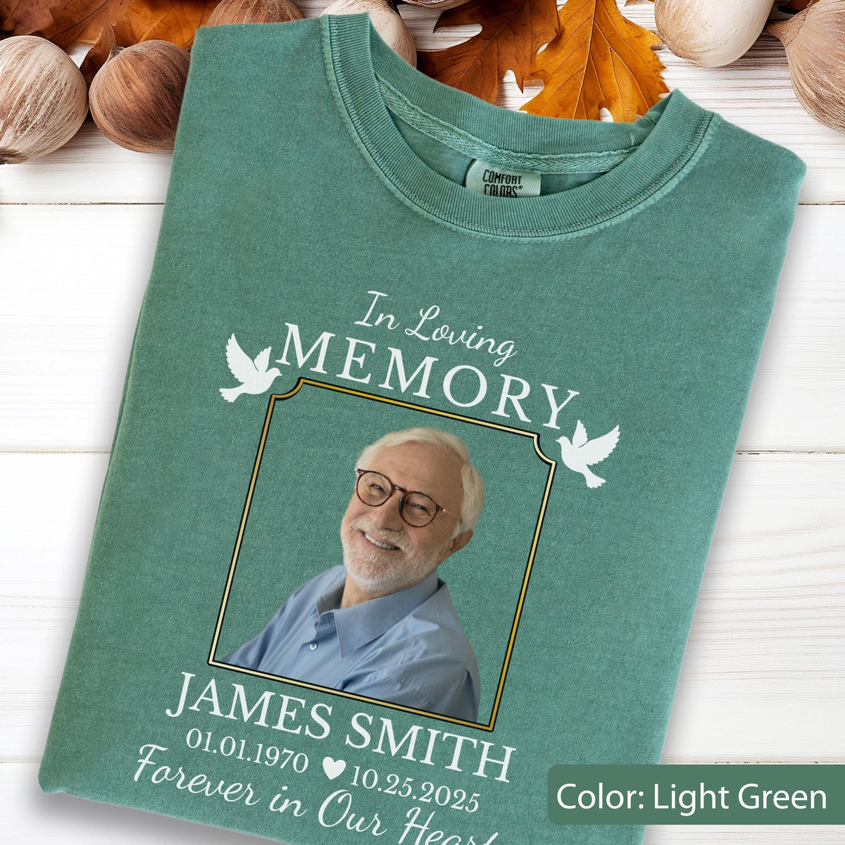 May include: A light green t-shirt featuring a memorial graphic with the text In Loving Memory, a photograph of a smiling person with glasses, the name James Smith, the dates 01.01.1970 and 10.25.2025, and two white dove illustrations.