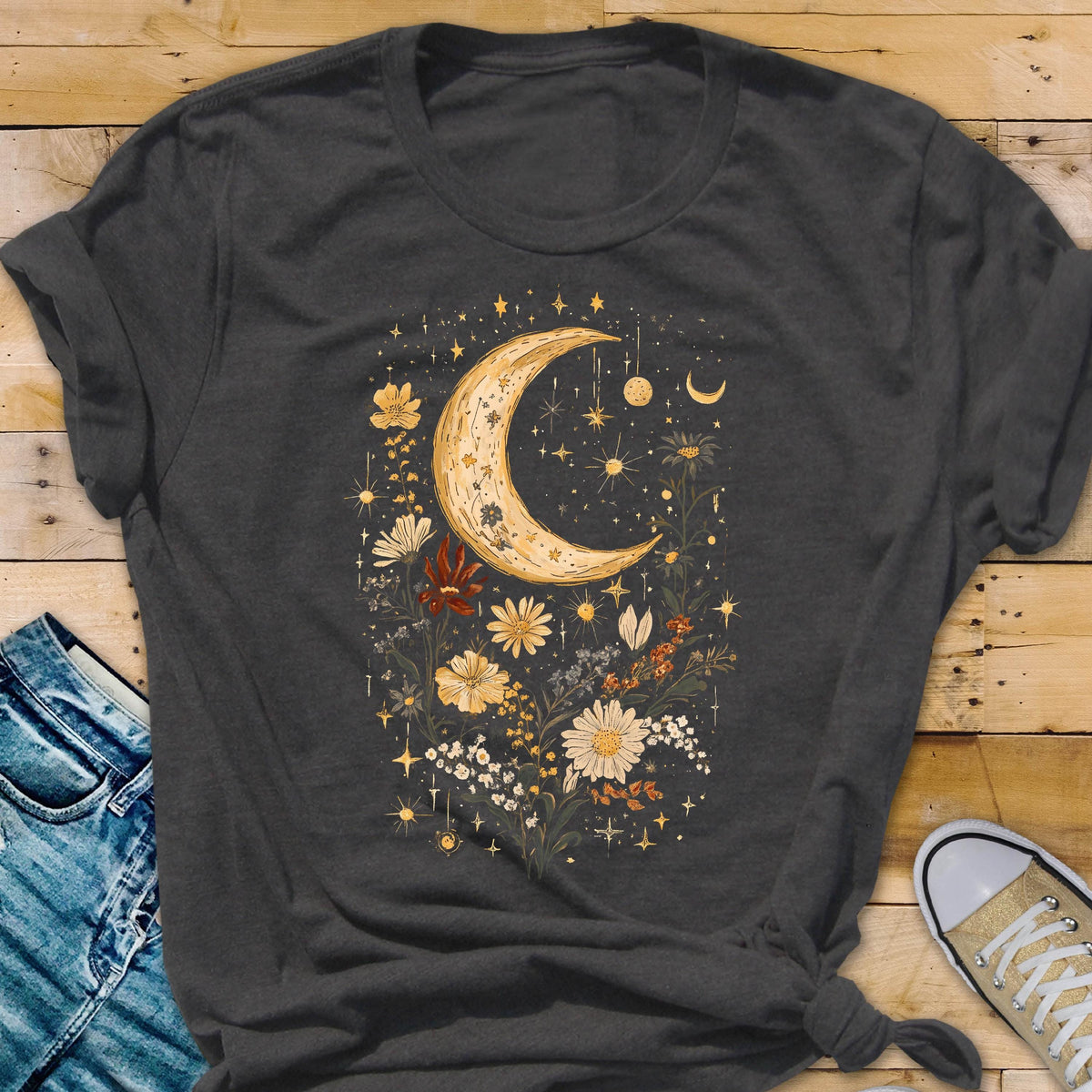 May include: Dark gray t-shirt featuring a crescent moon and floral design. The design includes a large, textured crescent moon, stars, and various flowers in shades of yellow, white, and red. The shirt is made of a soft material.