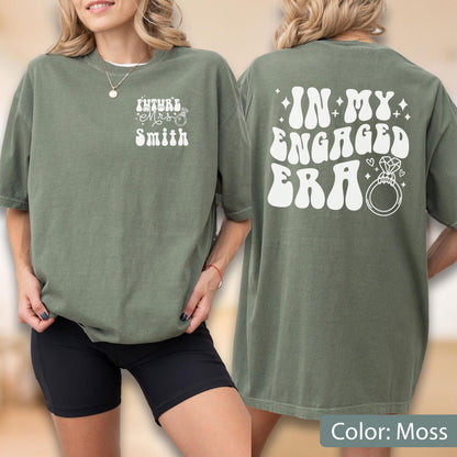 May include: A moss green short-sleeve t-shirt featuring white text. The front displays "FUTURE MRS SMITH" with a small ring icon. The back features the large white text "IN MY ENGAGED ERA" accompanied by a diamond ring graphic and small star accents.