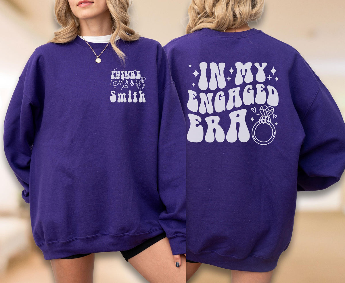 May include: A deep purple long-sleeve sweatshirt featuring white screen-printed text. The front displays "Future Mrs. Smith" with a small ring icon. The back features the large text "IN MY ENGAGED ERA" accompanied by a graphic of a diamond ring.