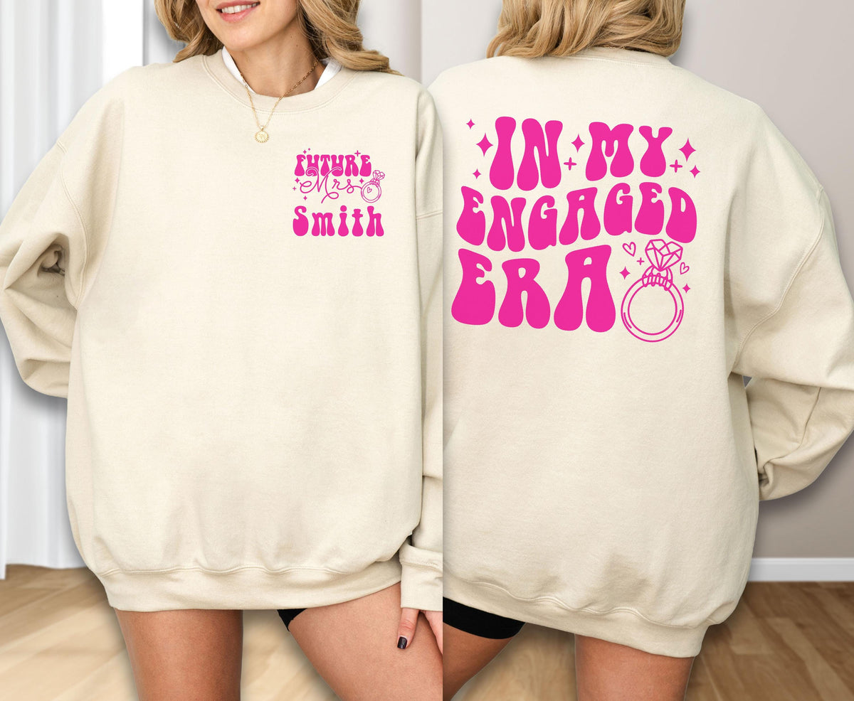 May include: A beige crewneck sweatshirt featuring hot pink text and graphics. The front left chest displays the text "Future Mrs. Smith" with a small ring icon. The back features the large text "IN MY ENGAGED ERA" with a diamond ring graphic.