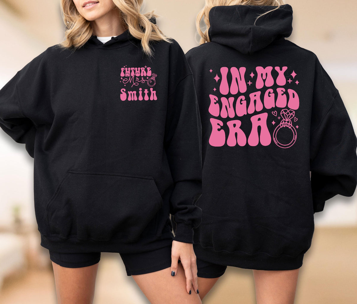 May include: A black hooded sweatshirt featuring pink text and graphics. The front left chest displays "FUTURE MRS SMITH" with a ring icon. The back features the text "IN MY ENGAGED ERA" in a large, bold, wavy font alongside a diamond ring graphic.